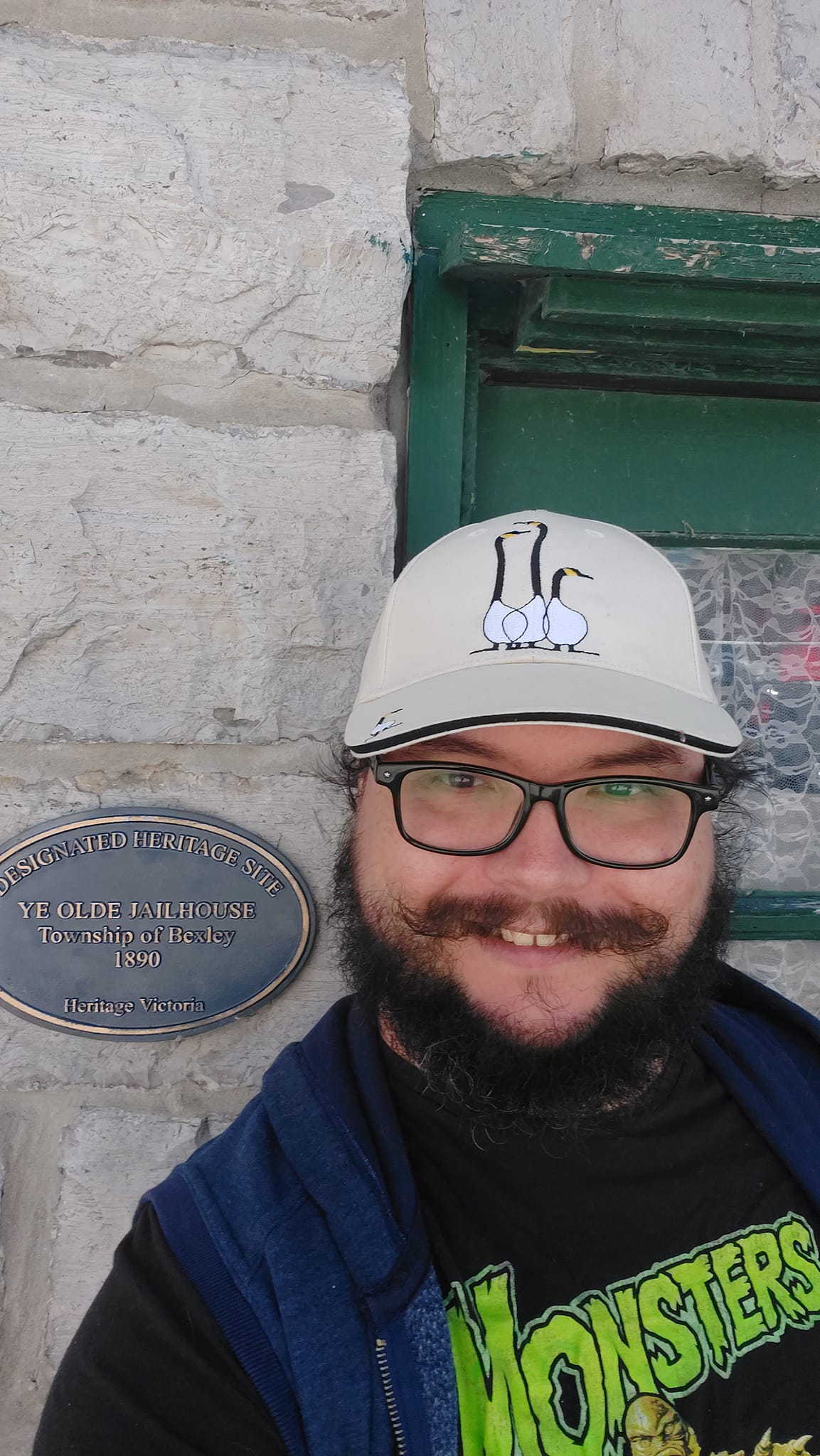 A man with short hair and bushy facial hair stands in front of a stone building with a plaque that reads "Designated Heritage Site, Ye Olde Jailhouse, Township of Bexley, 1890, Heritage Victoria"